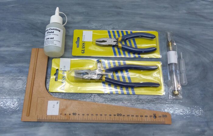 glass cutting kit with pliers, square rule, cutting tool and fluid on grey streaky glass background.