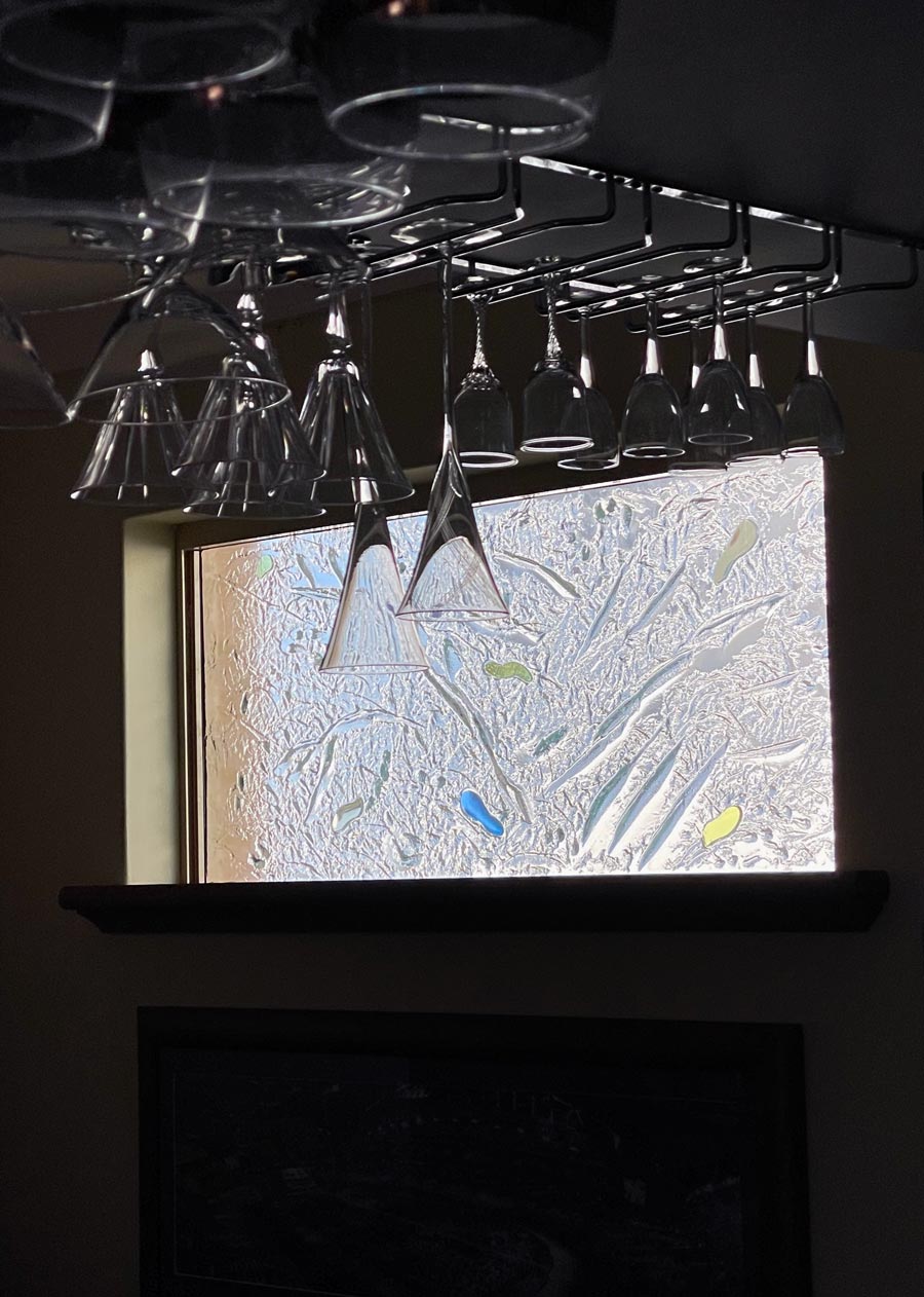 glasses hanging from bar in foreground and kiln formed window with laminated coloured pieces in background