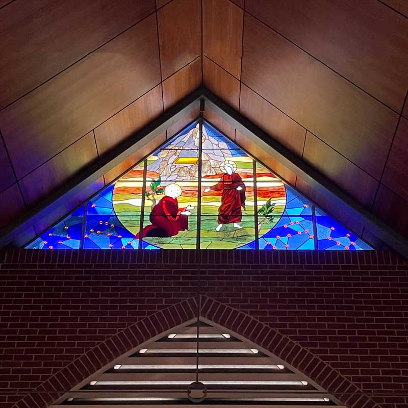 triangular stained glass window of Jesus and disciple in fields surrounded by church brickwork